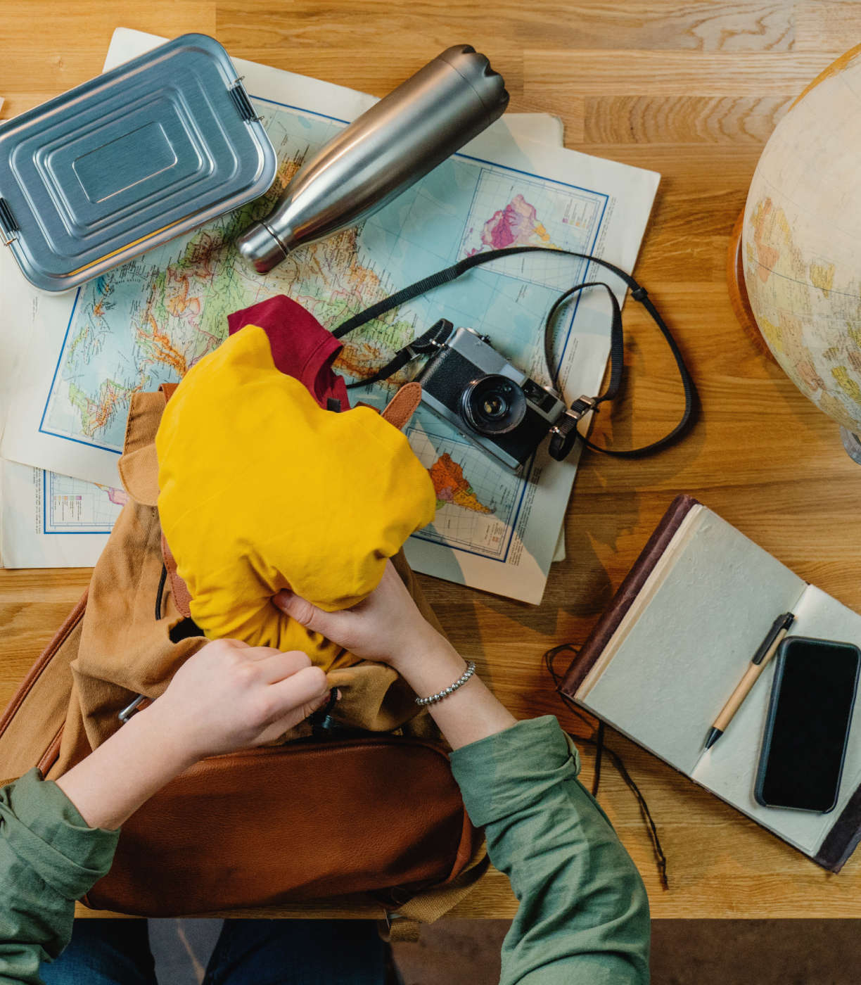 Travelers items. From top to bottom: Metal box and metal water bottle and camera on top of a map, a globe, clothes behind placed inside a bag, an open notebook with a pen and a phone laying on top.